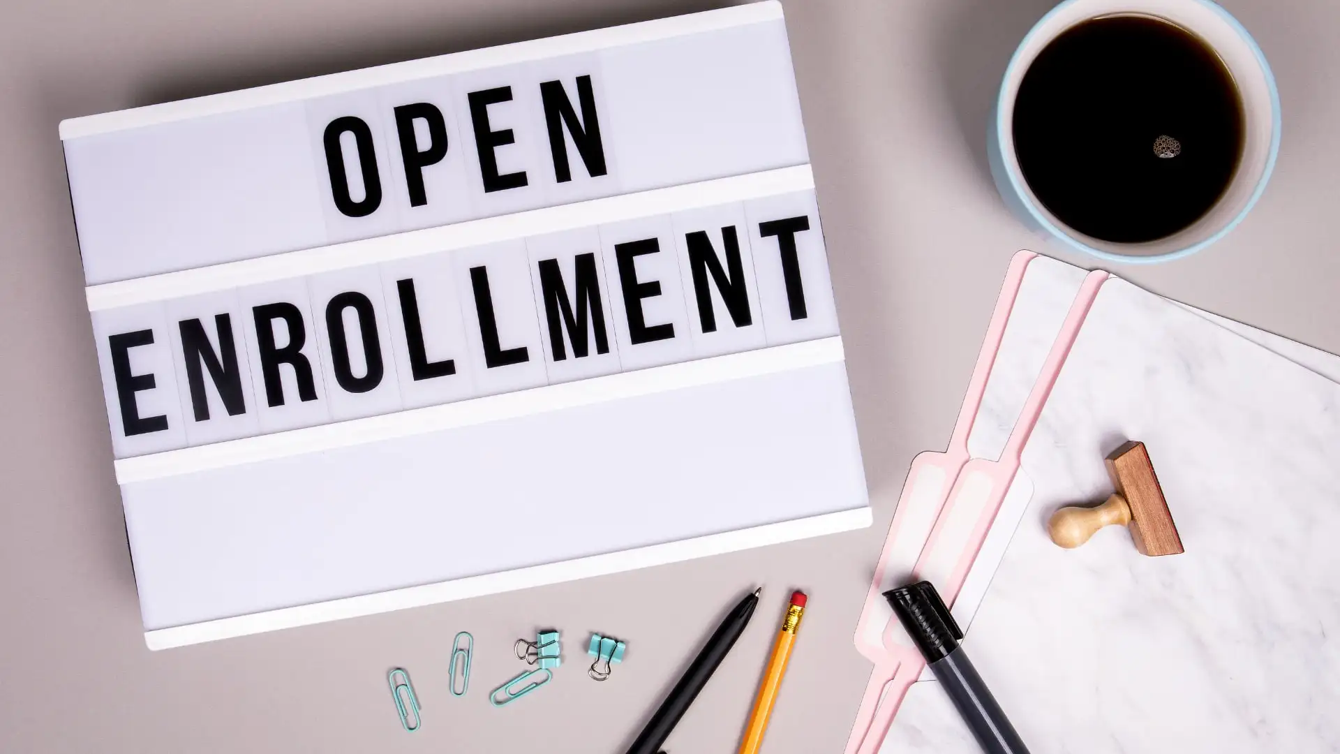 Caja de luz con las palabras Open Enrollment sobre un escritorio con café, carpetas y bolígrafos